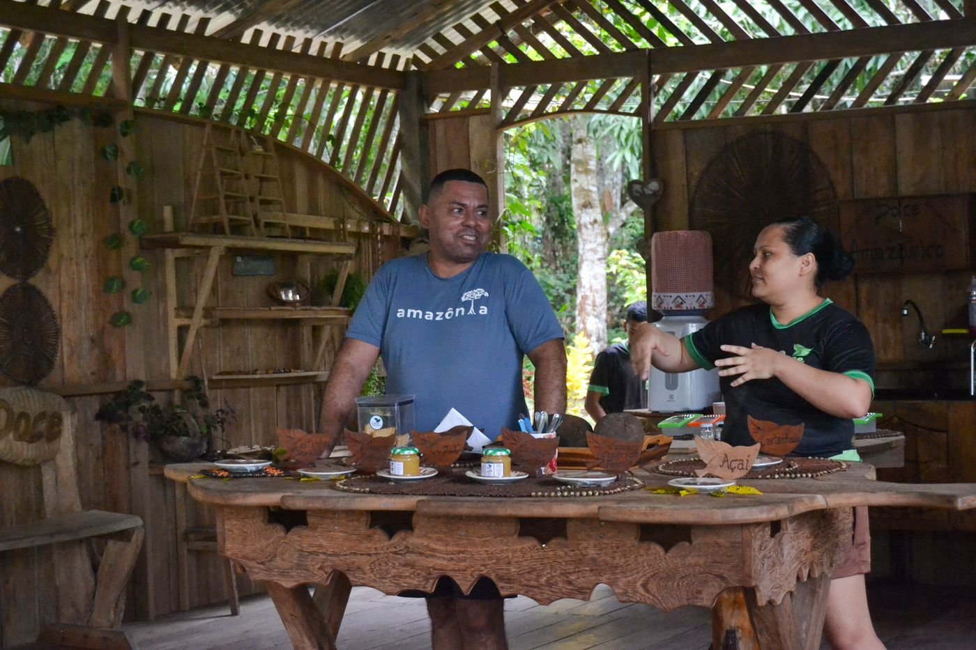 Empreendedores comunitários apresentam doces e produtos amazônicos sobre uma mesa de madeira, em um espaço aberto para a floresta.