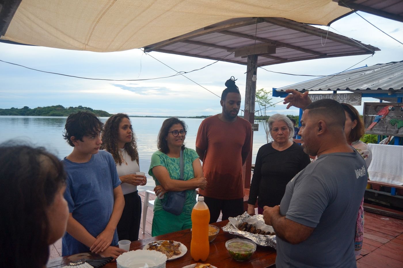 Participantes da imersão reunidos em um flutuante à beira do rio, ouvindo um morador da comunidade ao lado de uma mesa com comida local.